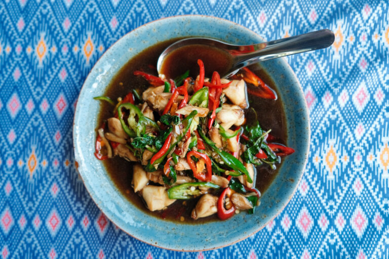 Fotografía gastronómica cenital de comida callejera tailandesa: Pad Kra Pao Gai, pollo salteado con albahaca sagrada y chiles en un cuenco azul sobre un mantel típico de Tailandia